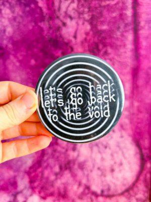 Duża gotycka przypinka w kolorze czarno białym z napisem let's go back to the void i spiralą w tle.