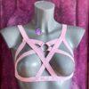 Różowa elastyczna uprząż na ciało typu body harness z krzyżującymi się paskami na biuście i metalowym kółkiem O-ring na środku, prezentowana na manekinie – pastel pink body harness, kawaii lingerie, strappy bra harness, fetish harness, Valentine lingerie, alternative underwear.